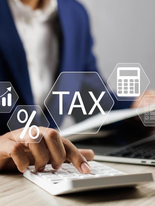 A professional in a blue suit calculating taxes using a calculator at a desk with a laptop and digital icons of documents, percentage sign, and tax symbol floating above the desk, representing online banking services. The person is focused on calculating taxes.